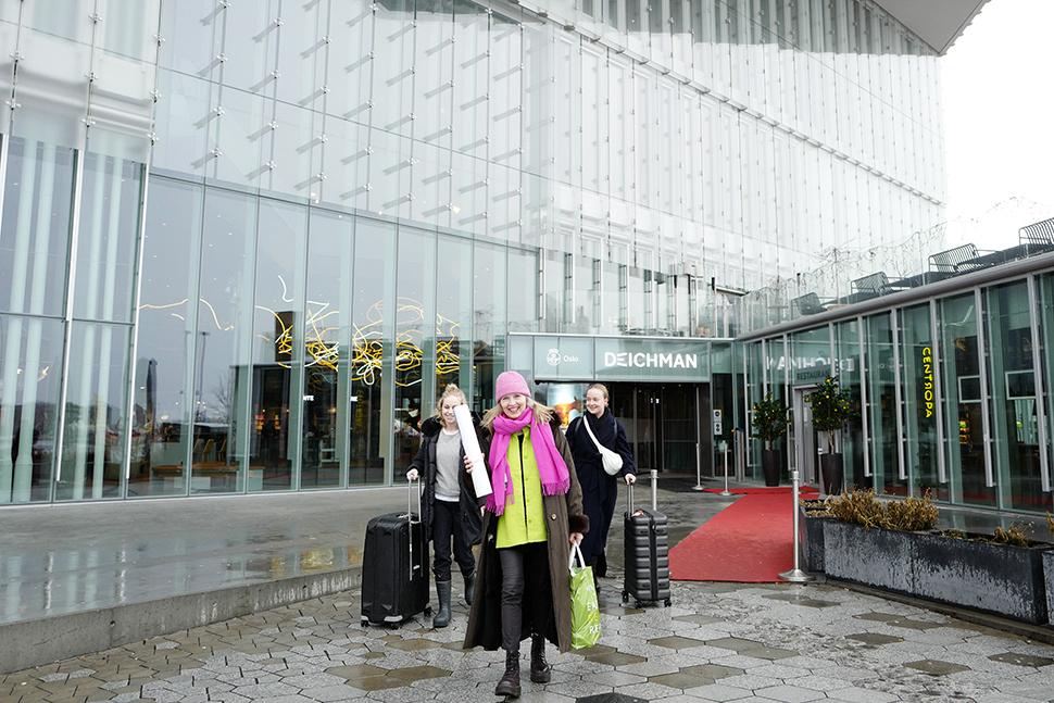 Tre smilende damer går med kofferter ut av glassbygning.